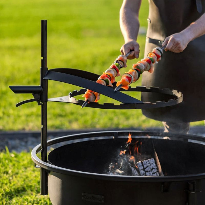 Man placerar grillspett på höjdjusterbart galler över eldfat från Cook King Bandito.