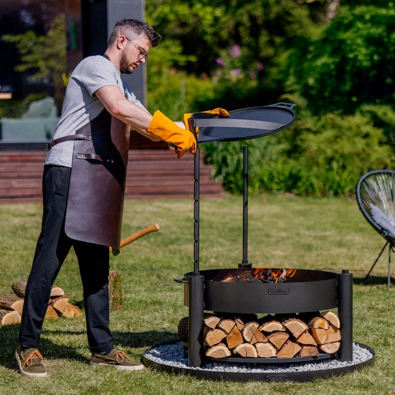 Man som justerar höjden på grillgallret över ett brinnande MONTANA X eldfat i trädgårdsmiljö