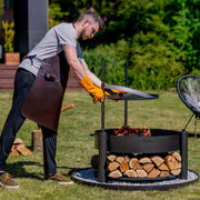Man placerar en stekhäll i råstål på MONTANA X eldskål från Cook King, redo för matlagning över öppen eld i trädgården.