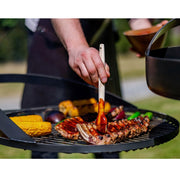 Grillgaller fyllt med kött och grönsaker över eld, där någon penslar sås på revbensspjäll