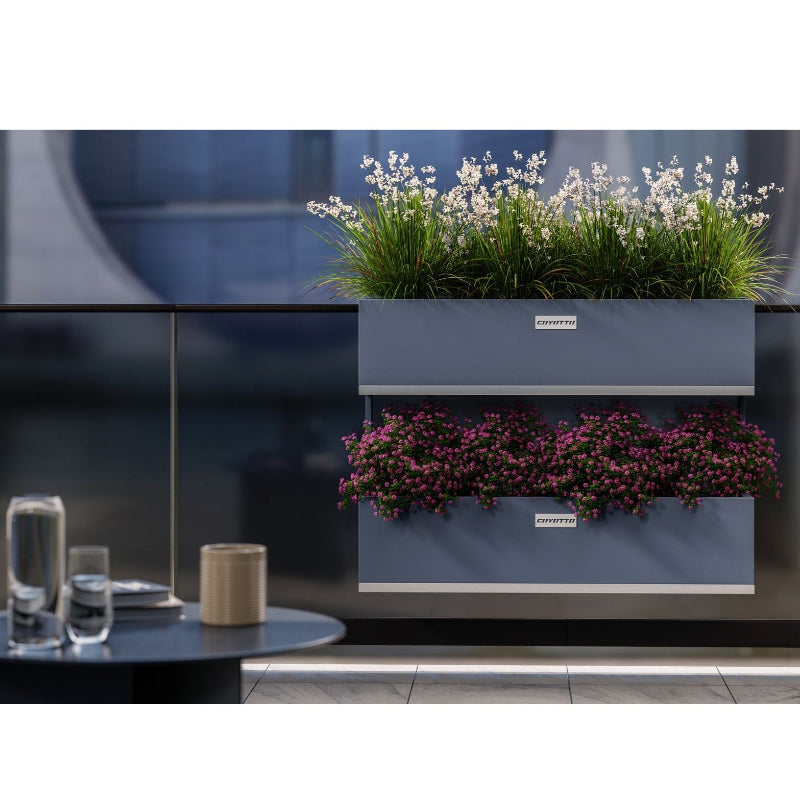 Två våningar av Coyotto balkonglådor monterade på glasräcke, fyllda med grönskande växter och färgstarka blommor – stilrent och funktionellt för urbana utemiljöer.