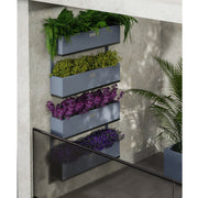 Fyra väggmonterade Coyotto planteringskärl i stål, fyllda med färgstarka växter och placerade på en balkongvägg med grönskande omgivning.